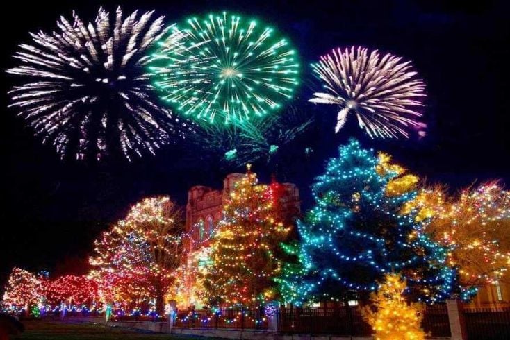 A building surrounded by Christmas trees with Christmas lights, and a fireworks display in the background