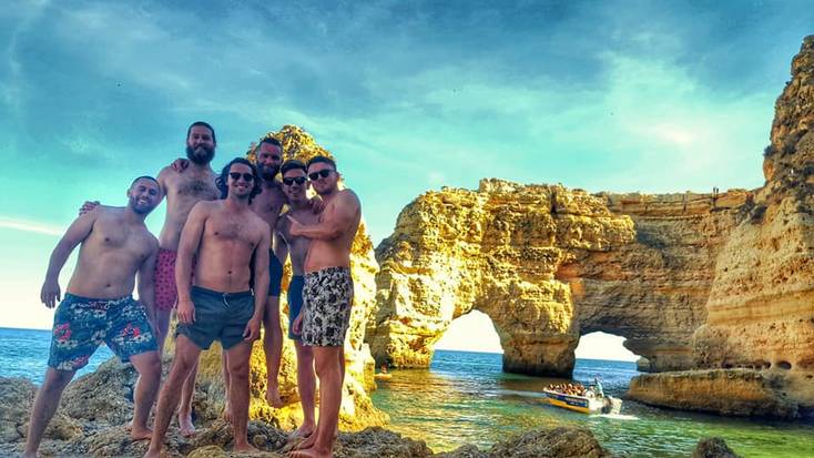 A group of glampers exploring the rocks and coves at Praia da Marinha, one of the best beaches in Portugal, while a boat tour of the caves takes place in the background