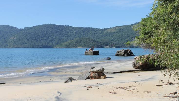 Brazil has some of the best beaches in the world, such as this beach on Ilha Grande, Brazil, surrounded by forest and travelers standing on a rock in the water.